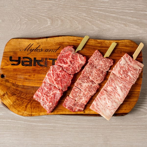 Three raw wagyu beef skewers on a wooden cutting board with "Myles and Jun Yakitori" engraved.