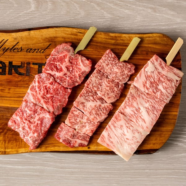 Three skewers of raw, marbled wagyu beef cuts on a wooden serving board.